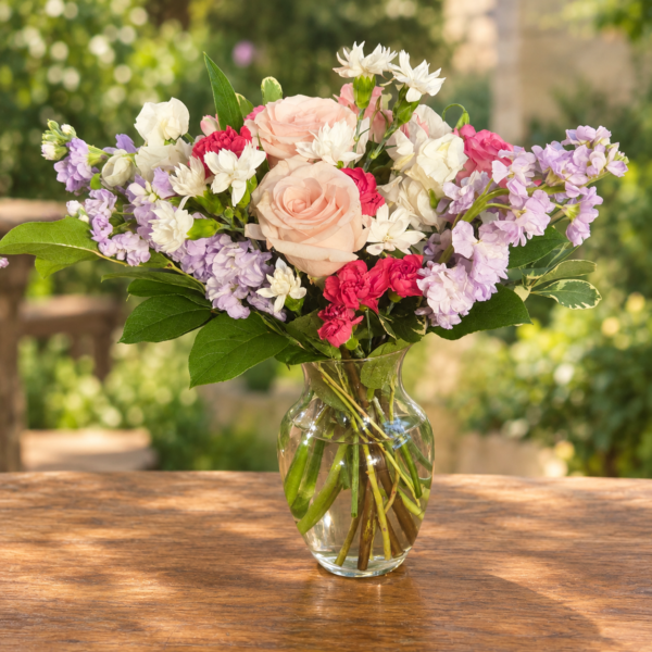 Exotic Floral Bouquet featuring blush pink roses, lavender stock, bright pink carnations, white accent blooms, and greenery arranged in a clear glass vase.