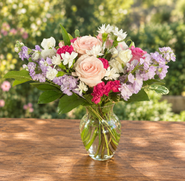 Exotic Floral Bouquet featuring blush pink roses, lavender stock, bright pink carnations, white accent blooms, and greenery arranged in a clear glass vase.