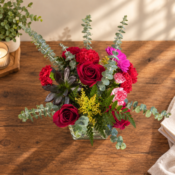 Red roses, a dark plum succulent, yellow solidago, and arching eucalyptus stems arranged in a clear glass cube vase.